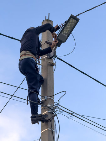 A man is working on a light pole. He is wearing a black shirt and blue pants. He is on a ladder
