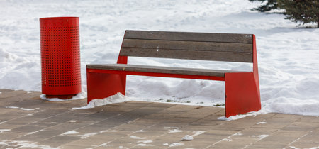A red bench sits on a sidewalk next to a red trash can. The bench is empty. The snow is piled up on...