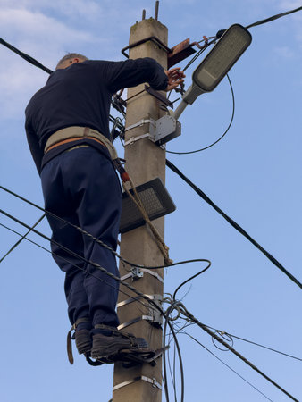 A man is working on a power pole. He is wearing a black shirt and blue pants. He is holding a light...