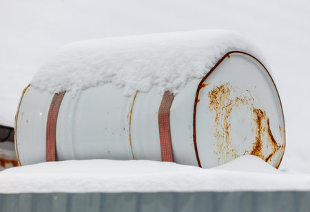 A rusty barrel covered in snow. The barrel is white and has a red strap. The barrel is sitting on top of snowの写真素材