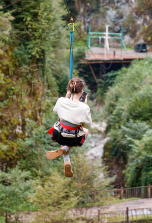 A woman is hanging from a zip line. She is wearing a white shirt and black shorts. She is holding a cell phone in her handの写真素材