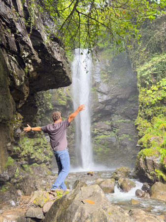 A man is standing in front of a waterfall. He is wearing a shirt and blue jeansの写真素材