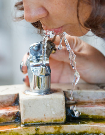 A woman is drinking water from a fountain. The water is cold and clear. The fountain is located on a sidewalkの写真素材