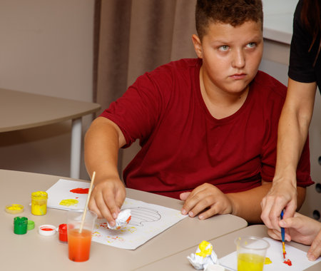 A boy is painting a picture with a brush. A woman is helping him. There are many different colors of...