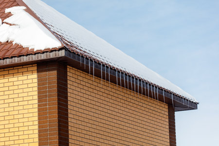 A brick building with a snow covered roof. The roof has icicles hanging from it. The building is...