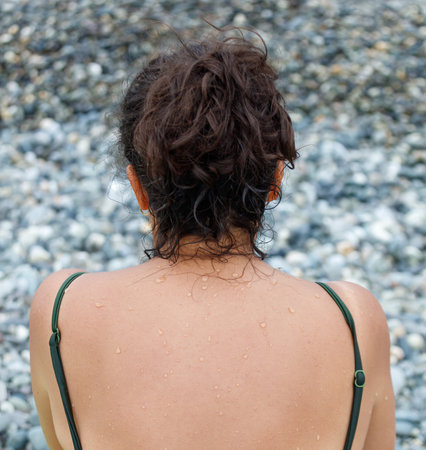 A woman with wet hair and a green top. She is standing on a rocky beach. The rocks are scattered...