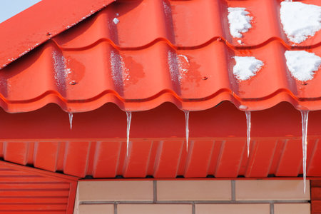 The roof of a building is covered in icicles, which are melting due to the warm weather. The icicles are hanging from the roof and are dripping water onto the ground below. The roof is red in colorの写真素材