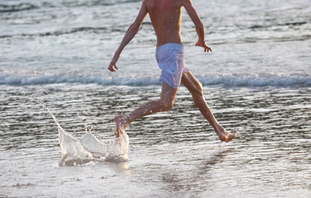 A man is running in the ocean. He is barefoot. The water is splashing. The man is wearing white shortsの写真素材