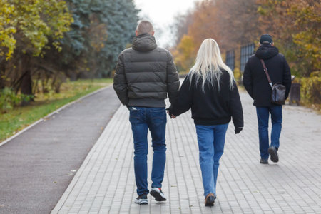 A man and a woman are walking on a sidewalk. The man is wearing a green jacket and jeans. A third...