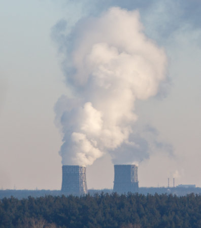 Two large power plants are emitting smoke into the sky. The smoke is white and billowing. The power...