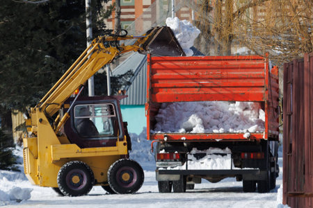 A yellow machine is lifting a pile of snow. The snow is being dumped into an orange truck