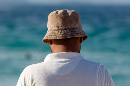 A man wearing a brown hat and a white shirt is standing on the beach. The hat is a straw hat with a...