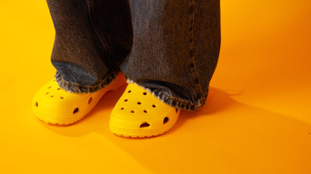 Close-up of person wearing yellow clogs and denim jeans. Feet stand on bright orange floor. Warm,...