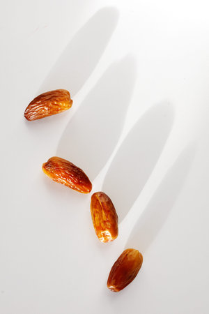 Four dried dates arranged diagonally on white surface. Soft shadows fall beside each fruit. Smooth,...