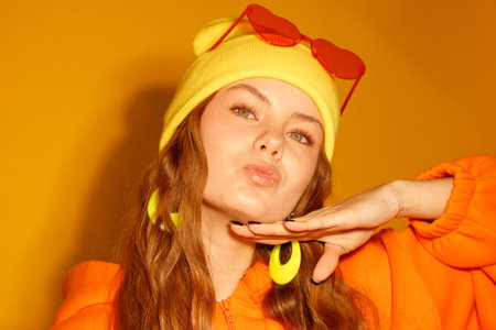 Young woman poses with hand near lips. Wears yellow beanie and orange hoodie. Background is solid...