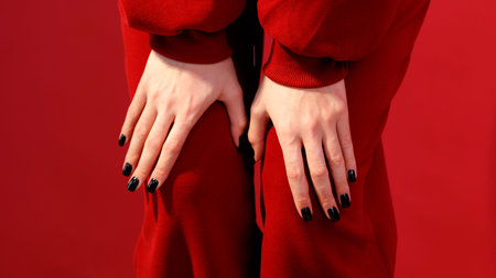 Close-up of woman's hands with black nail polish. Hands rest on red fabric. Background is solid red....
