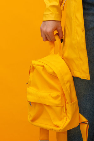 Person carries bright yellow backpack against matching yellow background. Hand grips backpack strap with dark nail polish. Person wears yellow jacket and blue jeans