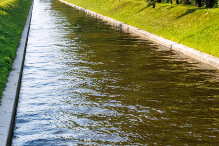Clear water flows gently through a canal. Concrete edges line both sides of the waterway. Lush green grass grows beside the canal walls. Sunlight glints softly on the rippling surface.