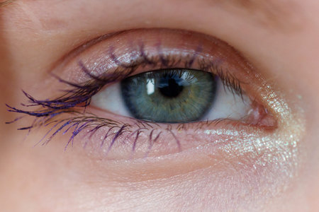 Close-up of human eye with green iris and dark pupil. Eyelashes frame the eye, slightly curled upward. Skin around eye shows natural texture and subtle shine. Iris has gentle green hue with soft