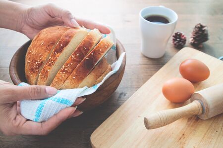 Woman hand holding loaf of homemade bread with sesame on wood table background with eggs and cup of coffeeの写真素材