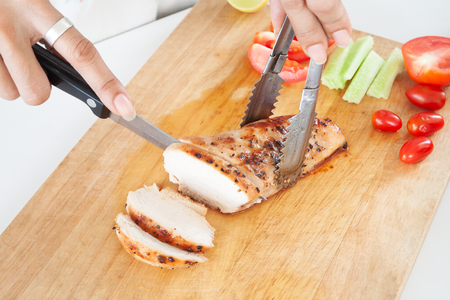Woman's hand cutting grilled chicken breast with tomatoes and cucumber on cutting board, Close upの写真素材