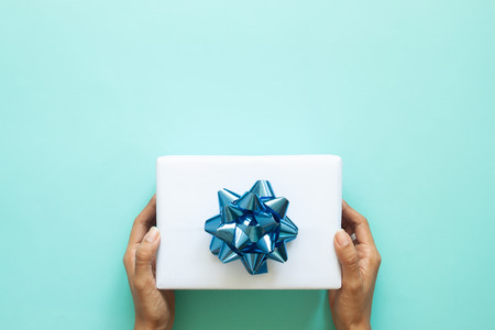 Woman's hands holding white gift box with blue ribbon on pastel color background