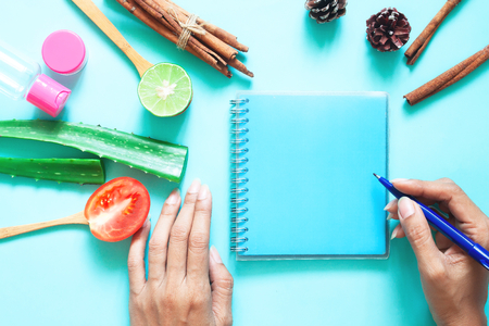 Creative flat lay of skincare and healthcare concept. Woman's hands with empty notebook, tomato, aloe vera, lime and cosmetic containers on pastel backgroundの写真素材