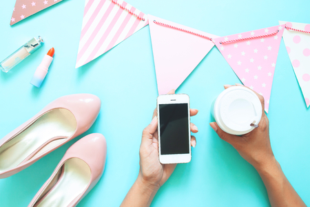 Woman's hand holding smart phone and coffee cup with pastel colour bunting and accessories on colour backgroundの写真素材