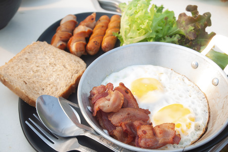 Fried eggs, bacon and sausage on pan, sliced bread and vegetable on a plate. concept breakfast.の写真素材