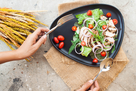 Creative flat lay of salad with grilled chicken, onions and tomatoes on black plate with woman's handsの写真素材
