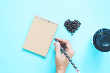Creative flat lay of heart shape coffee beans and woman hand holding pencil with notebook on blue colour backgroundの写真素材