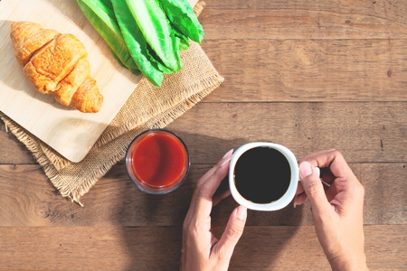 Top view woman hands holding black coffee cup, tomato juice, croissant and vegetable on rustic wooden tableの写真素材