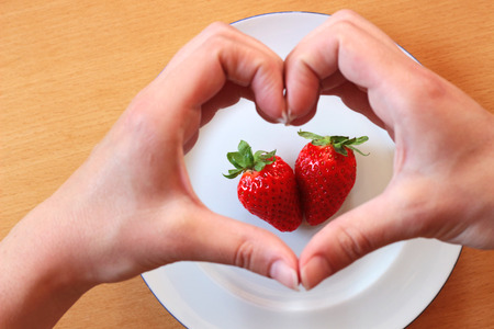 hands forming a heart over two strawberries lying on a white plate on a tableの写真素材
