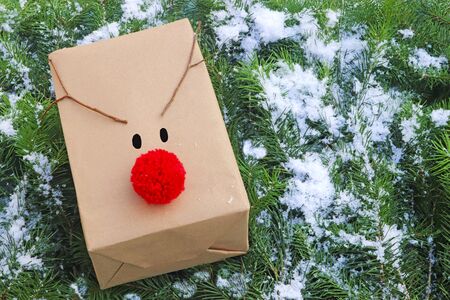 christmas background with a gift parcel decorated with a red reindeer nose and antlers, lying on snow covered pine branchesの写真素材