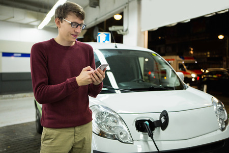 Man is holding a phone. His electric car charged at the charging stationの写真素材