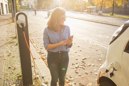 Young woman is standing near the electric car and holding smartphone. The rental car is charging at the charging station for electric vehicles. Car sharing.の写真素材