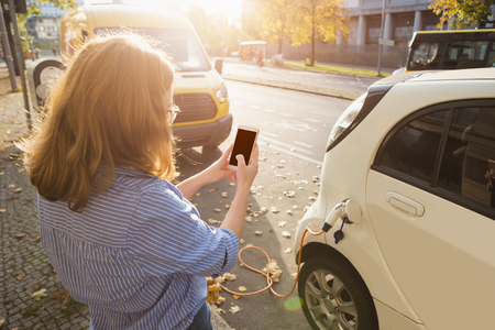 Young woman is standing near the electric car and holding smartphone. The rental car is charging at the charging station for electric vehicles. Car sharing.の写真素材