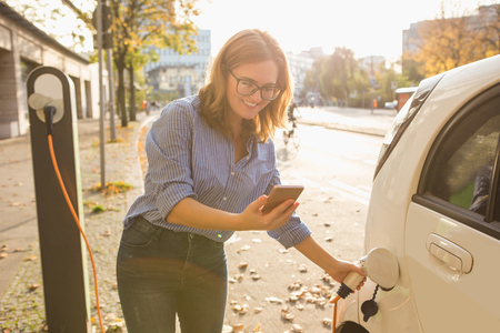 Young woman is standing near the electric car and holding smartphone. The rental car is charging at the charging station for electric vehicles. Car sharing.の写真素材