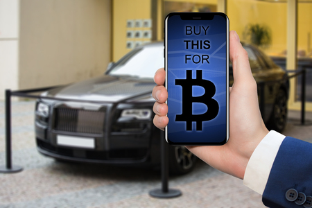 Hand of a man with a phone against the background of a luxury car. On the screen is the inscription "Buy this for bitcoin". The symbol of wealth made on the crypto currencyの写真素材