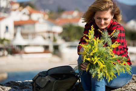 Portrait of a woman holding a bouquet of mimosa flowers. The girl on the beach. A woman is dressed in a red shirt and blue jeans.の写真素材