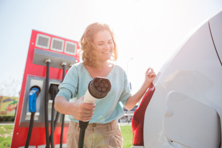 A woman stands at the charging station and holds a plug of the charger for an electric carの写真素材