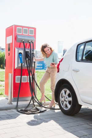 A woman stands at the charging station and holds a plug of the charger for an electric carの写真素材