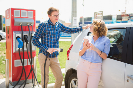 Smiling man and woman on the charging station for electric cars. A man is charging a car, a woman is standing with a phone.の写真素材