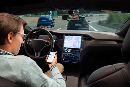 A man reads news online in a smartphone while his car is driven by an autopilot. Self driving vehicle conceptの写真素材