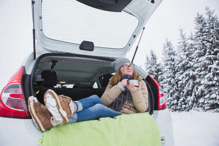 Woman sits in the trunk of the car and holds a cup of hot tea in her hands.の写真素材