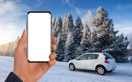Hand with a phone on the background of a car behind a winter road. Blank screen, you can add your contentの写真素材