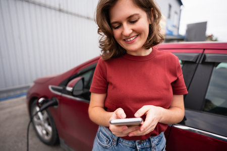 Woman with smartphone charging red electric carの写真素材