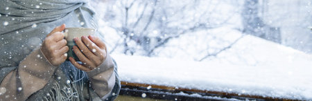 A woman in a warm hat and blanket holds a mug with a hot drink in her hands. Winter landscape with...
