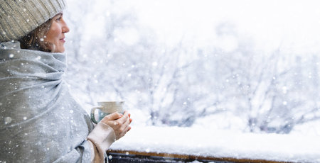 A woman in a warm hat and blanket holds a mug with a hot drink in her hands. Winter landscape with snowfall in the background.の写真素材