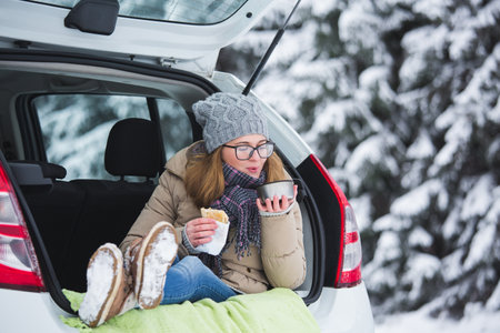 Woman in woolen hat sits in the trunk of the car and holds a cup of hot tea in her hands.の写真素材
